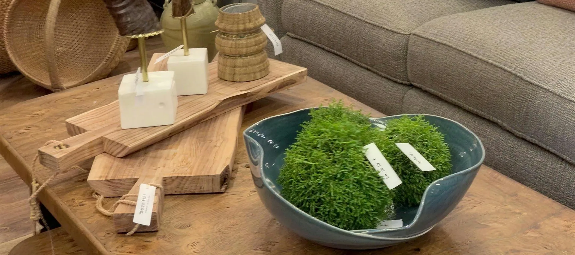 A burl wood coffee table with serving boards, a blue bowl of moss and a taupe sofa behind it